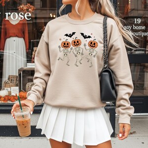May include: Beige sweatshirt featuring a Halloween design with three skeleton figures with pumpkin heads, bats, and stars. The text "amy rose" is visible in the upper left corner. The sweatshirt is paired with a white pleated skirt.