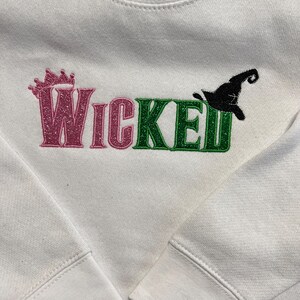 May include: White sweatshirt with the word "Wicked" in pink and green glitter lettering. A pink crown is above the word "Wicked" and a black witch hat is next to the word "Wicked".