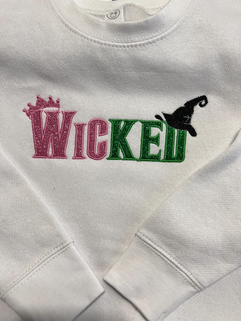 May include: White sweatshirt with the word "Wicked" in pink and green glitter lettering. A pink crown is above the word "Wicked" and a black witch hat is next to the word "Wicked".