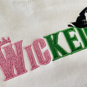 May include: White sweatshirt with the word "WICKED" embroidered in pink and green. The "W" features a crown, and a black witch hat sits atop the "E". The letters have a glittery texture.