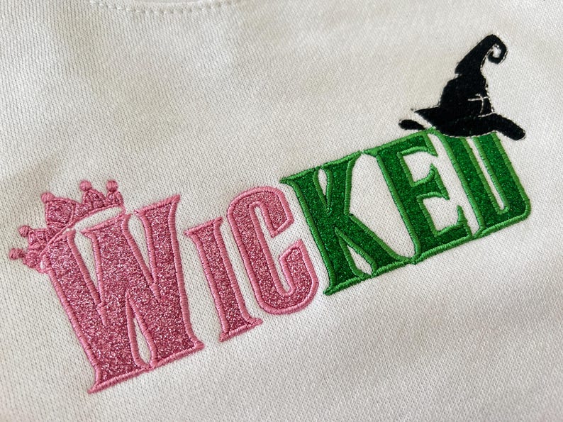 May include: White sweatshirt with the word "WICKED" embroidered in pink and green. The "W" features a crown, and a black witch hat sits atop the "E". The letters have a glittery texture.