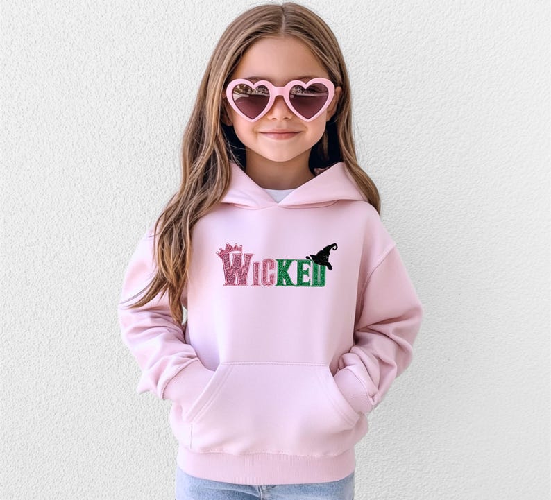 May include: A light pink hooded sweatshirt with the word "Wicked" in glittery pink and green letters, and a black witch hat graphic. The sweatshirt has a front pocket and is being worn by a young person wearing heart-shaped sunglasses.