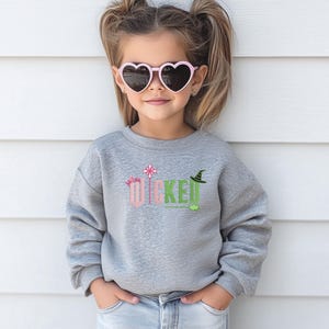May include: A young girl wearing a gray sweatshirt with the word "WICKED" in pink and green letters. The letters are decorated with a crown, a wand, and a witch's hat. She is wearing pink heart-shaped sunglasses.