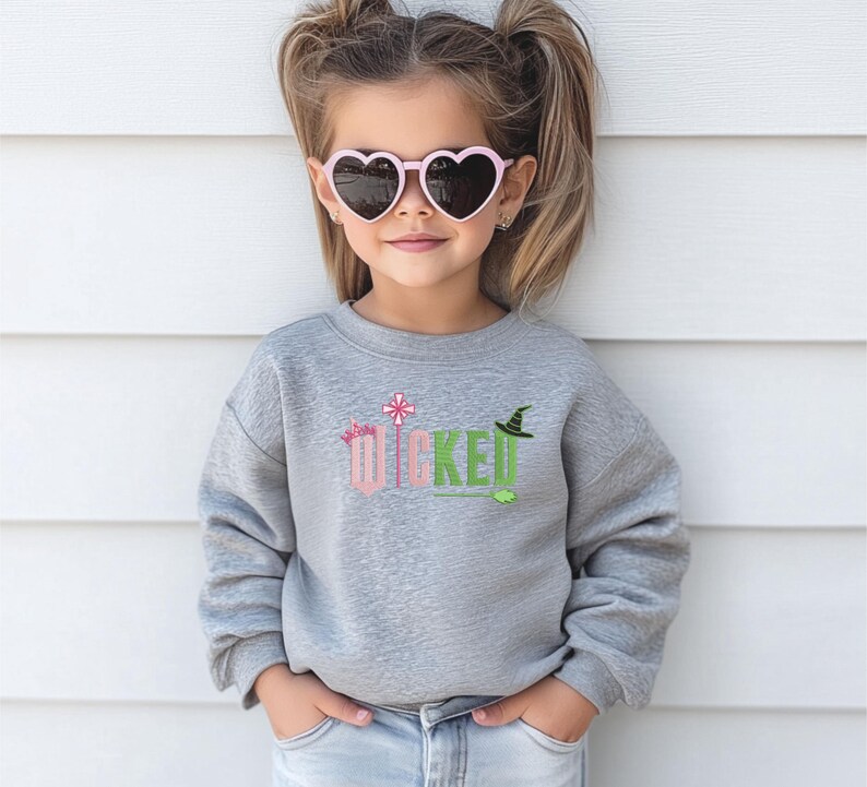 May include: A young girl wearing a gray sweatshirt with the word "WICKED" in pink and green letters. The letters are decorated with a crown, a wand, and a witch's hat. She is wearing pink heart-shaped sunglasses.