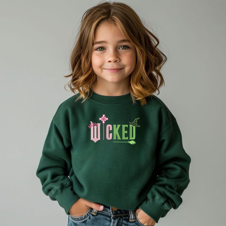 May include: A dark green sweatshirt with the word 'WICKED' printed on the front in pink and green letters. The word is surrounded by small pink and green graphic elements, including a crown, a wand, a witch's hat, and a broom.