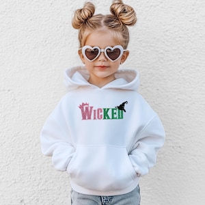 May include: A white hooded sweatshirt with the word "Wicked" in pink and green glitter lettering. A black witch hat graphic is on the right side of the word. The sweatshirt has a front pocket and is worn by a child wearing heart-shaped sunglasses.