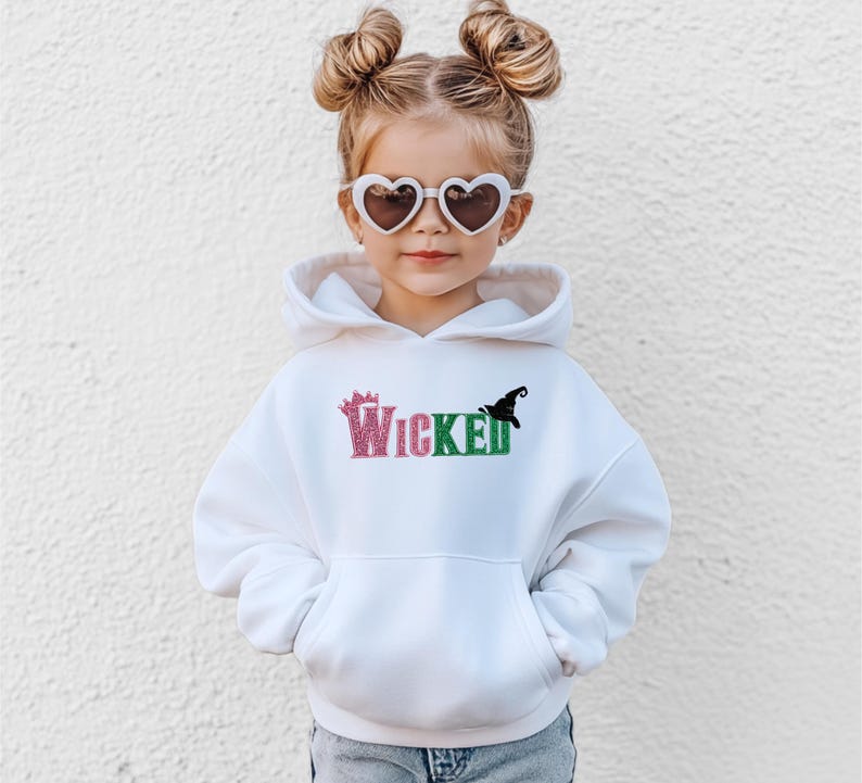 May include: A white hooded sweatshirt with the word "Wicked" in pink and green glitter lettering. A black witch hat graphic is on the right side of the word. The sweatshirt has a front pocket and is worn by a child wearing heart-shaped sunglasses.