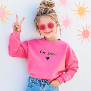 May include: A young person wearing a bright pink sweatshirt with the words "for good" and a green heart. The person is also wearing pink sunglasses and blue jeans. The background features a white wall with pink and yellow sunburst designs.