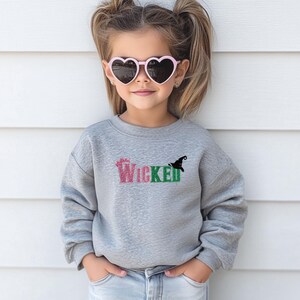 May include: A young person wearing a gray sweatshirt with the word "Wicked" in pink and green glitter lettering. A black witch hat graphic is next to the word. The person is also wearing pink heart-shaped sunglasses.