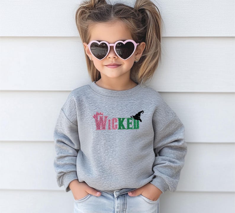 May include: A young person wearing a gray sweatshirt with the word "Wicked" in pink and green glitter lettering. A black witch hat graphic is next to the word. The person is also wearing pink heart-shaped sunglasses.