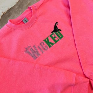 May include: Bright pink sweatshirt with the word "Wicked" in glittery pink and green lettering, along with a black witch hat graphic. A crew neck sweatshirt, perfect for Halloween or a witch-themed event.