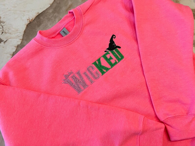 May include: Bright pink sweatshirt with the word "Wicked" in glittery pink and green lettering, along with a black witch hat graphic. A crew neck sweatshirt, perfect for Halloween or a witch-themed event.