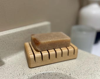May include: A light brown bar of soap rests on a wooden soap dish. The soap dish is made of light-colored wood with a slatted design. The soap has a rectangular shape and a smooth surface. The items are on a speckled countertop.
