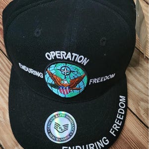 May include: Black baseball cap with the words "OPERATION ENDURING FREEDOM" embroidered in white. The cap features a patch with an eagle and globe design. A holographic sticker reads "GENUINE RAPID DOMINANCE MERCHANDISE."