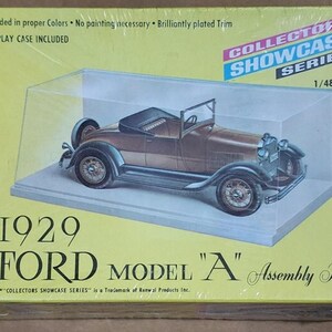 Może przedstawiać: Zestaw do montażu Forda Model A z 1929 roku w gablotce. Model samochodu jest brązowo-srebrny, z czarnymi oponami i kołem zapasowym z tyłu. Pudełko jest żółte z napisem "Collectors Showcase Series".