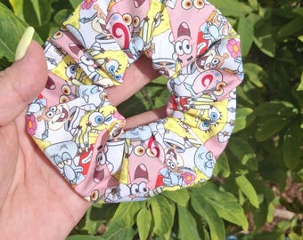 May include: A fabric hair scrunchie featuring a repeating cartoon print of various characters including a yellow sponge, a pink starfish, and a light blue octopus against a white background with scattered pink and yellow floral accents.