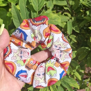 May include: A fabric hair scrunchie featuring a repeating pattern of various Muppet characters including Kermit the Frog, Miss Piggy, Fozzie Bear, and Animal on a white background. The scrunchie has a gathered, ruffled texture and a circular shape.