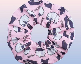 May include: Pink fabric scrunchie with a repeating pattern of a cartoon character with white hair and black eyes. The character is wearing a black mask and a black outfit. The text "呪術廻戦" is repeated throughout the pattern.