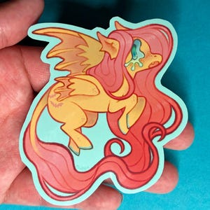 May include: A sticker depicting a cartoon pony in yellow and pink hues. The pony has wings and long, flowing hair. The sticker has a light blue border and is held in a hand.