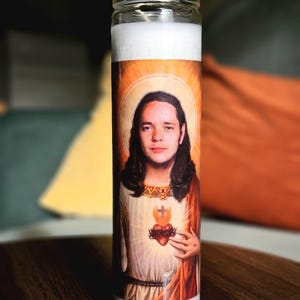 以下が含まれることがあります： 長い髪の人物、光輪、いばらの冠の心臓の肖像画が描かれた透明なガラスの祈りのろうそく。 ろうそくは白いワックスの内側とオレンジと黄色のラベルが付いています。