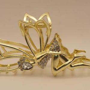 Könnte beinhalten: Haarspange aus goldfarbenem Metall mit floralem Design. Der Clip hat einen Mittelteil, der mit kleinen, funkelnden Strasssteinen und einem tropfenförmigen Juwel verziert ist. Das Design des Clips umfasst offene Blütenblätter und einen sicheren Verschluss.