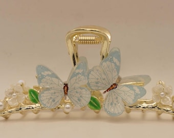 May include: Gold-coloured hair claw clip featuring two pale blue butterflies with detailed wing patterns. The clip is adorned with white floral accents, green leaves, and faux pearls, creating a nature-inspired design.