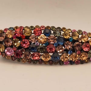 Op de afbeelding: Een ovale haarclip, versierd met een veelvoud aan kleurrijke strass-steentjes. De clip heeft een donkere metalen basis en is bedekt met een mozaïek van roze, blauwe, gele en bruine edelstenen, waardoor een sprankelend accessoire ontstaat.