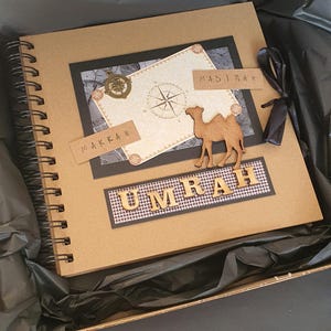 May include: A brown scrapbook with a black ribbon and a camel cutout. The cover has a compass, a map, and the words "Makkah" and "Madinah" written on it. The words "Umrah" are written in gold letters on a black and white checkered background.