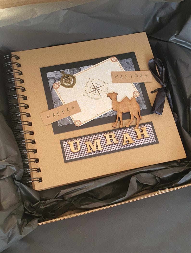 May include: A brown scrapbook with a black ribbon and a camel cutout. The cover has a compass, a map, and the words "Makkah" and "Madinah" written on it. The words "Umrah" are written in gold letters on a black and white checkered background.