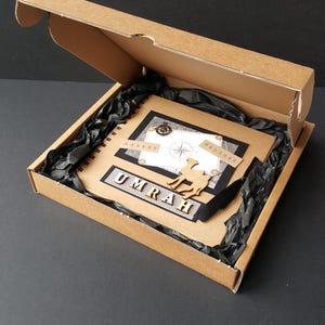 May include: A brown cardboard box with a black interior is open, revealing a black and white scrapbook with the word "UMRAH" in gold letters. The scrapbook has a compass, a camel, and the words "MAKKAH" and "MADINAH" on it.