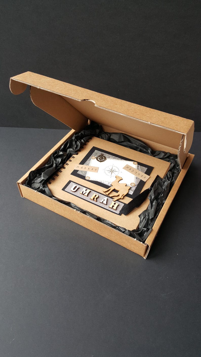 May include: A brown cardboard box with a black interior is open, revealing a black and white scrapbook with the word "UMRAH" in gold letters. The scrapbook has a compass, a camel, and the words "MAKKAH" and "MADINAH" on it.