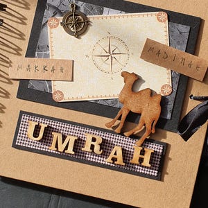 May include: A brown scrapbook with a black ribbon, a compass, and a wooden camel. The scrapbook has the words "Makkah" and "Madinah" on it. The words "Umrah" are spelled out in wooden letters.