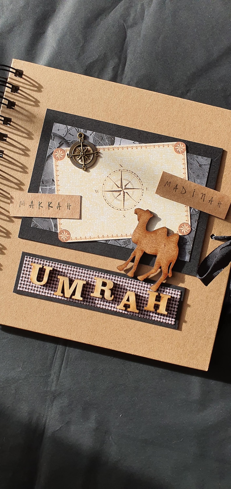 May include: A brown scrapbook with a black ribbon, a compass, and a wooden camel. The scrapbook has the words "Makkah" and "Madinah" on it. The words "Umrah" are spelled out in wooden letters.