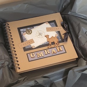 May include: A brown scrapbook with a black and white compass design and the words "Makkah" and "Madinah" on it. A wooden camel figurine is on the front of the book. The book is titled "Umrah" in wooden letters.