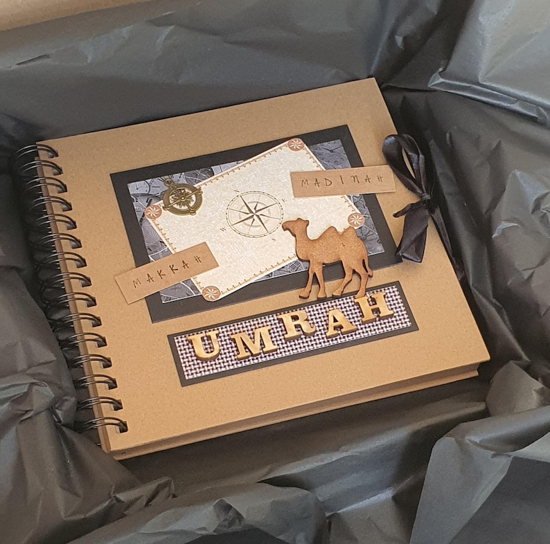 May include: A brown scrapbook with a black and white compass design and the words "Makkah" and "Madinah" on it. A wooden camel figurine is on the front of the book. The book is titled "Umrah" in wooden letters.