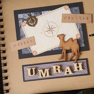 May include: A brown scrapbook with a black spine and a spiral binding. The cover features a black and white map with a compass, a camel cutout, and the words "Makkah", "Madinah", and "Umrah" in brown lettering.