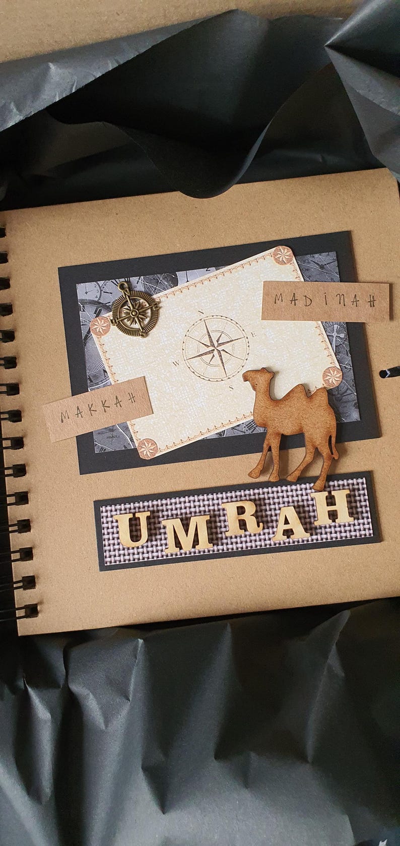 May include: A brown scrapbook with a black spine and a spiral binding. The cover features a black and white map with a compass, a camel cutout, and the words "Makkah", "Madinah", and "Umrah" in brown lettering.