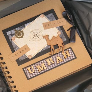 May include: A brown scrapbook with a black and white compass design. The cover has the words "Makkah" and "Madinah" written on it. The title of the scrapbook is "Umrah" and is made of wooden letters. The scrapbook is closed and has a black ribbon tied around it.
