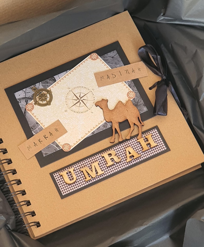 May include: A brown scrapbook with a black and white compass design. The cover has the words "Makkah" and "Madinah" written on it. The title of the scrapbook is "Umrah" and is made of wooden letters. The scrapbook is closed and has a black ribbon tied around it.