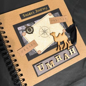 May include: A brown scrapbook with a black spiral binding. The cover has a gold title that says "Sajdas Journey" and a wooden camel cutout. The cover also has a compass and the words "Makkah" and "Madinah" written in gold.