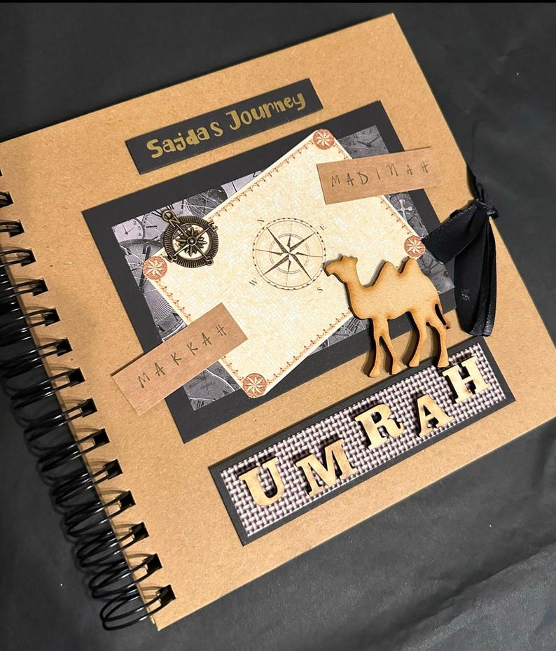 May include: A brown scrapbook with a black spiral binding. The cover has a gold title that says "Sajdas Journey" and a wooden camel cutout. The cover also has a compass and the words "Makkah" and "Madinah" written in gold.