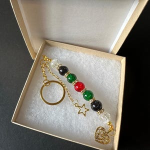 May include: A gold keychain with a beaded charm featuring black, green and red beads. The charm also includes a gold star and heart pendant.