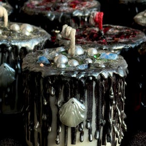 May include: A collection of decorative candles with a unique design. The candles are cylindrical, with a white base and black wax dripping down the sides. The tops are decorated with silver wax, faux pearls, and small blue and green embellishments.