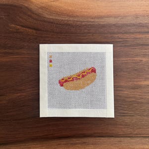 May include: A square piece of white needlepoint canvas featuring a cross-stitched hot dog in the center. The hot dog is tan with red ketchup and yellow mustard details. Three small colored squares are aligned vertically on the left side.
