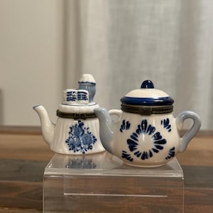 May include: Two small, decorative porcelain teapots. One is white with blue floral designs and a blue lid. The other is white with blue floral designs and a small blue structure on top. Both have gold-colored metal accents.