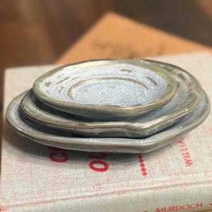 Puede incluir: Una pila de tres platos de cerámica de diferentes tamaños, cada uno con una forma orgánica única y un esmalte moteado de color azul grisáceo claro. Los platos descansan sobre un libro con el texto "Christ Is Our Victory".