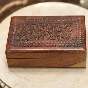 May include: A rectangular wooden box with a carved floral design on the lid. The box is a warm brown color and appears to be made of wood. The floral design is intricate and detailed, adding a decorative element to the box.