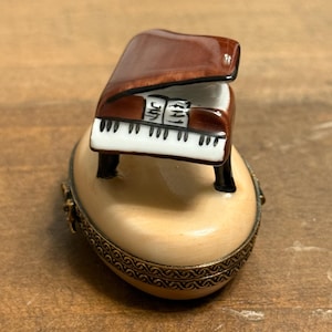Könnte beinhalten: Eine Miniatur-Flügel-Figur mit glänzend braunem Finish. Das Klavier sitzt auf einer cremefarbenen, eiförmigen Schmuckschatulle mit dekorativem goldfarbenem Besatz. Die Klaviertasten sind weiß und schwarz.