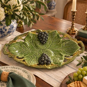 Puede incluir: Una bandeja de servicio ovalada de cerámica con un diseño de hojas verdes brillantes y dos racimos de uvas de color morado oscuro. La bandeja presenta un borde calado con un tono amarillento y asas integradas, mostrando una textura detallada.