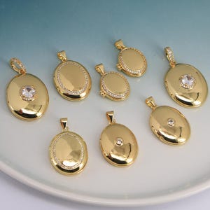 May include: A collection of gold-colored oval pendants. Some pendants feature a central clear gemstone, while others are adorned with a ring of small, clear stones. The pendants are displayed on a white surface.